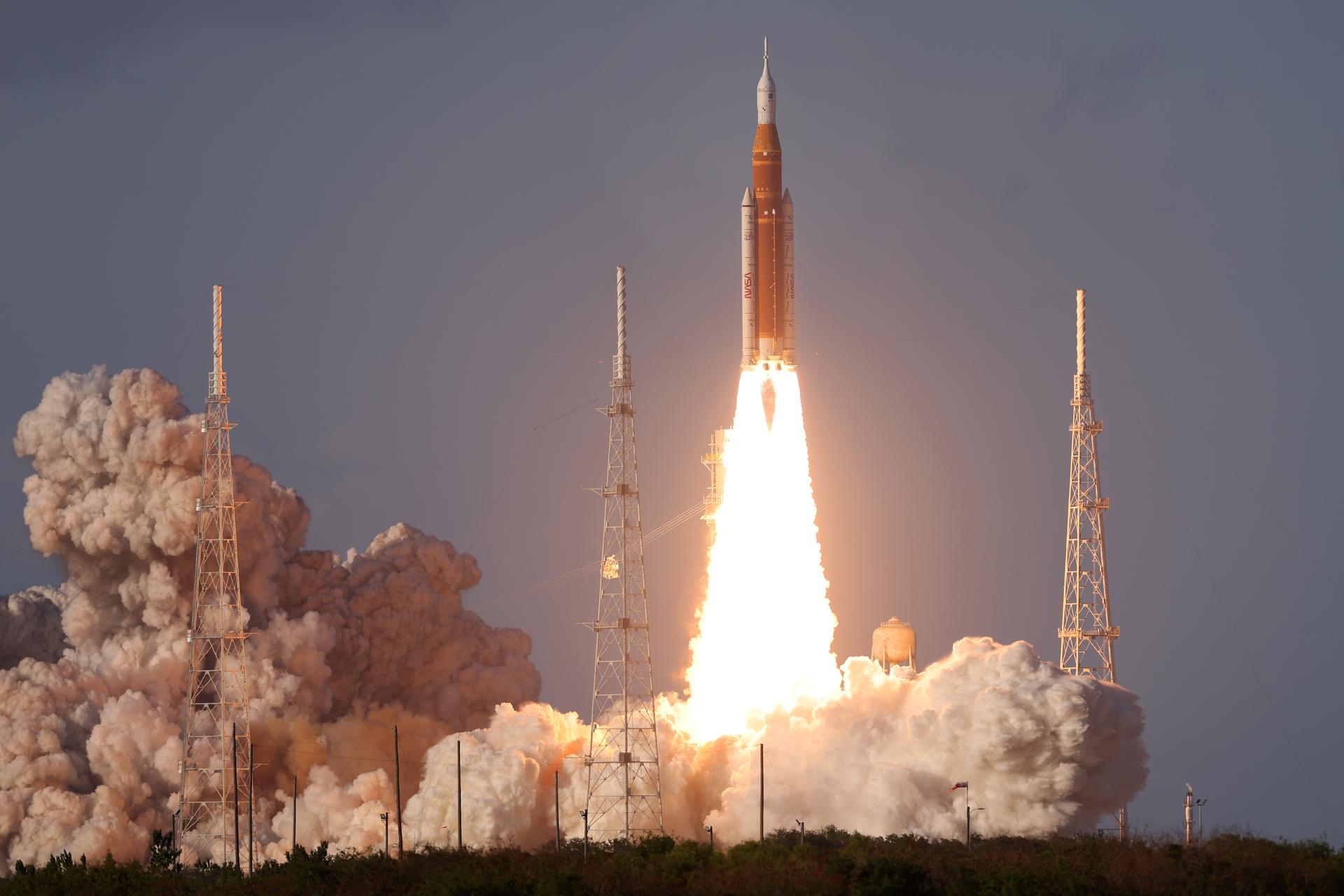 Artemis II SLS rocket climbing away from the launch pad at Kennedy Space Center, engine plume glowing bright orange against a grey sky.