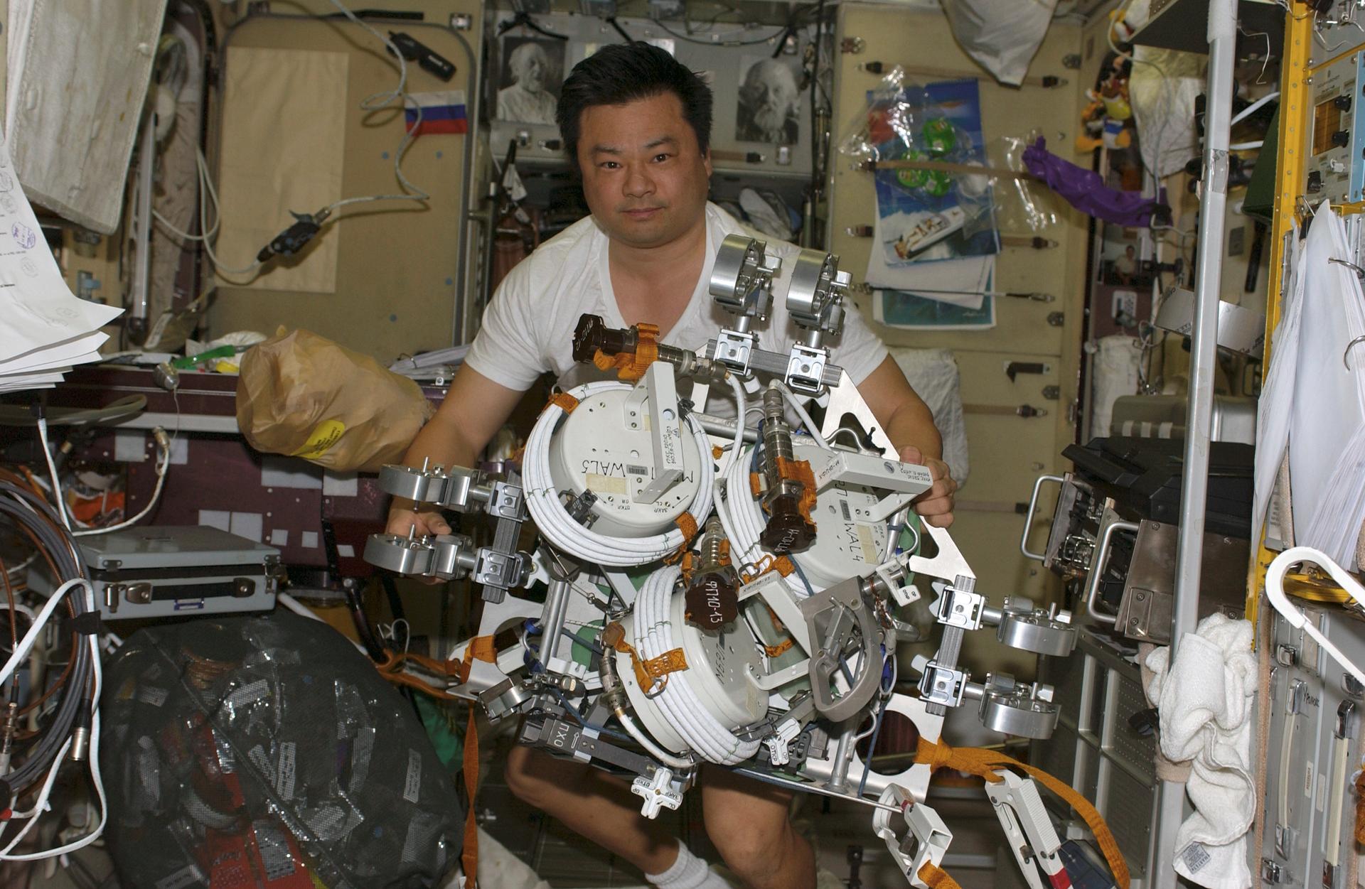 Astronaut aboard the International Space Station holding a partially assembled Extravehicular Mobility Unit backpack, surrounded by station equipment.