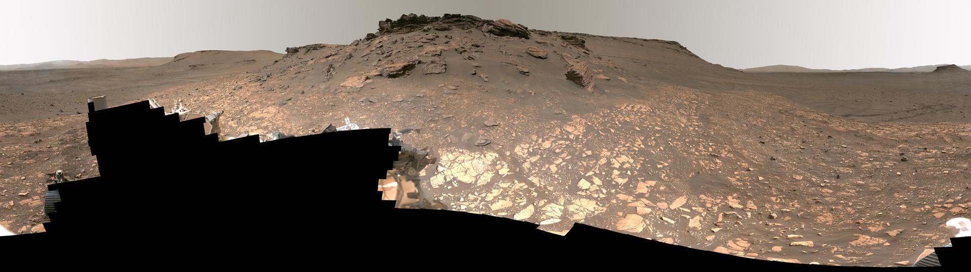 Panoramic view from the Perseverance rover of the layered delta terrain inside Jezero Crater, Mars, with the rover's own hardware visible in the foreground.