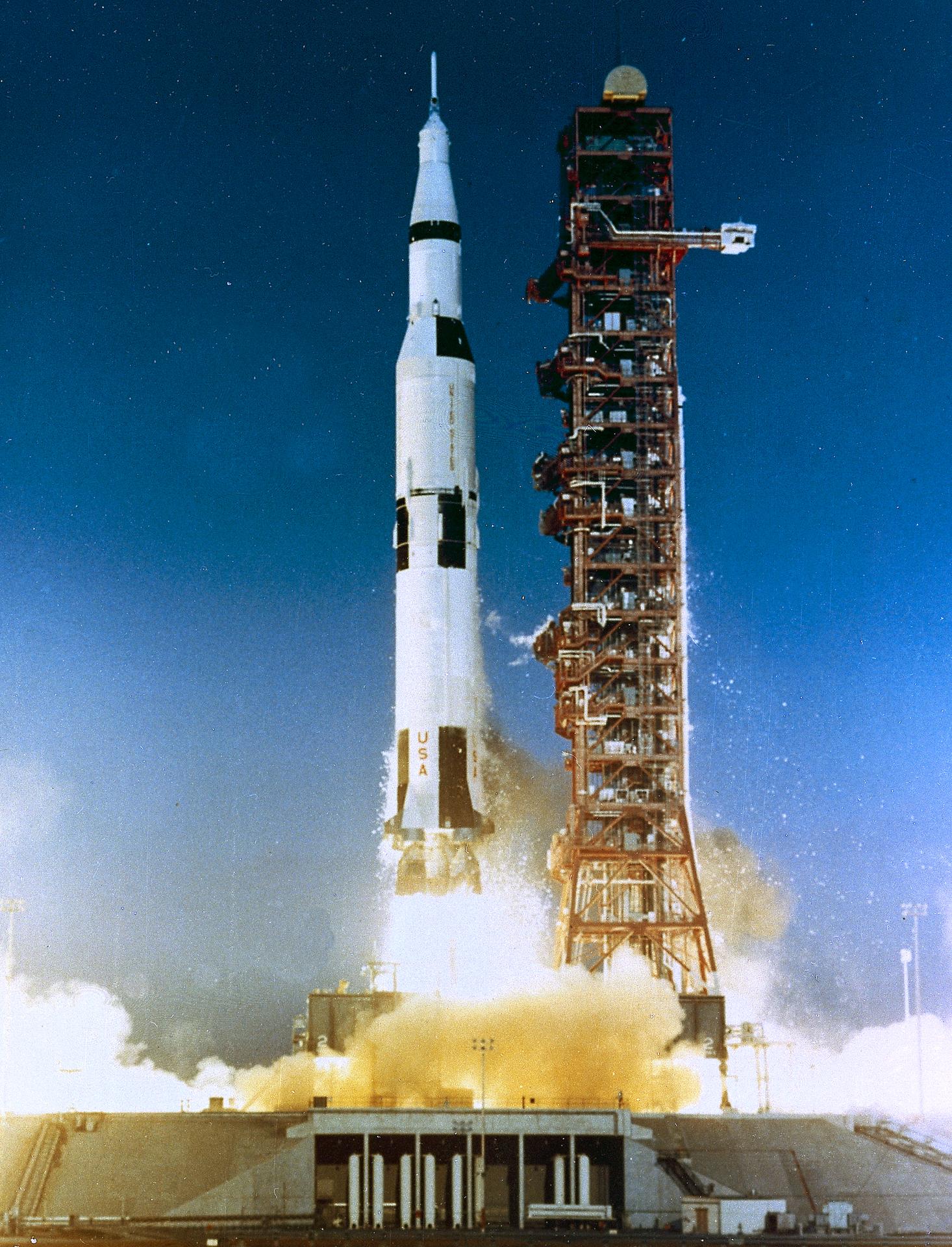 Apollo-era Saturn V rocket lifting off from Kennedy Space Center, exhaust plume billowing across the launch pad.